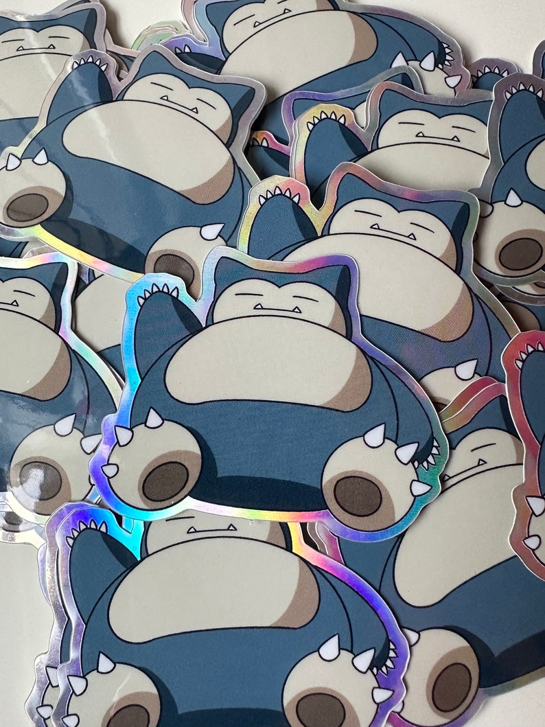 May include: A stack of holographic stickers featuring a cartoon Snorlax Pokemon character. The stickers are blue, grey, and white with a shiny, iridescent finish.