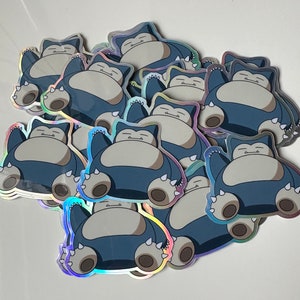 May include: A stack of holographic stickers featuring a cartoon Snorlax character. The stickers are blue, grey, and white.
