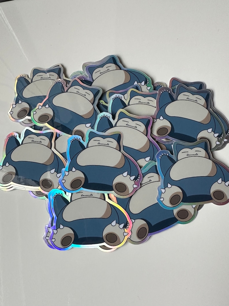 May include: A stack of holographic stickers featuring a cartoon Snorlax character. The stickers are blue, grey, and white.