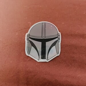 May include: A sticker featuring the Mandalorian helmet design. The helmet is primarily gray with black accents and a distinctive T-shaped visor. The sticker is on a reddish-brown surface.