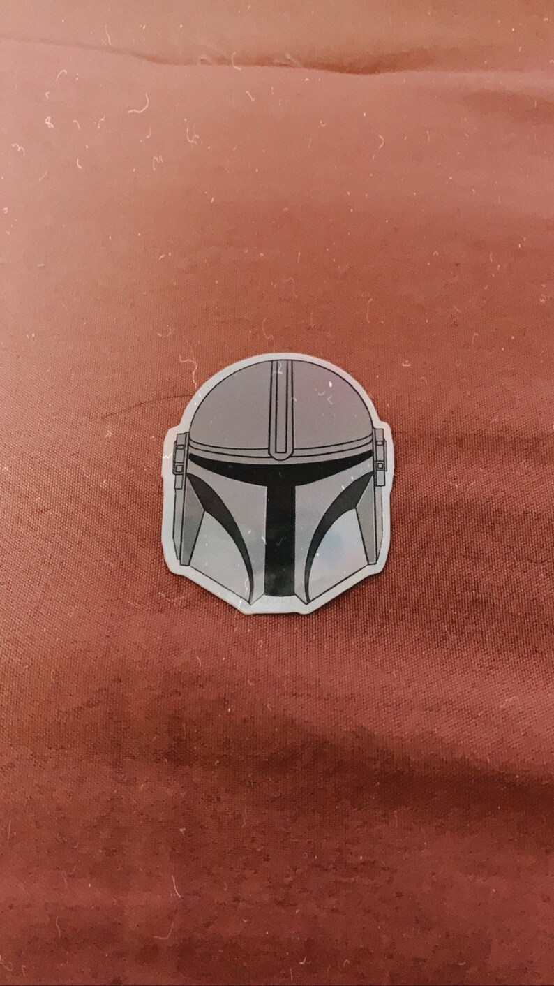May include: A sticker featuring the Mandalorian helmet design. The helmet is primarily gray with black accents and a distinctive T-shaped visor. The sticker is on a reddish-brown surface.