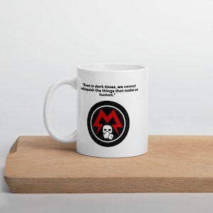 May include: White ceramic mug with a black and red graphic and the text "Even in dark times, we cannot relinquish the things that make us human." The mug has a curved handle and is set on a wooden surface.