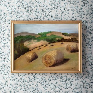 Può includere: Un dipinto incorniciato di un paesaggio rurale con balle di fieno in un campo. Il dipinto è su uno sfondo bianco con un motivo floreale blu.