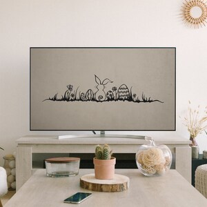 May include: A flat-screen TV displays a black and white Easter-themed graphic. The graphic features a bunny, Easter eggs, flowers, and grass. The TV is on a light-colored wooden table in a living room setting.
