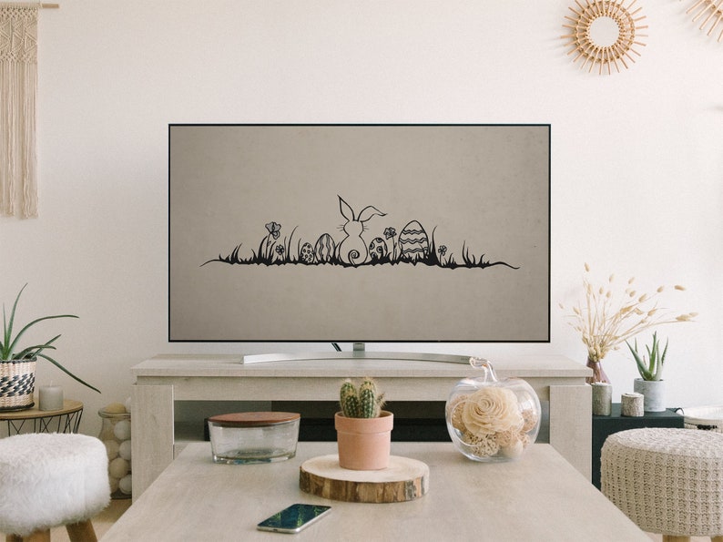 May include: A flat-screen TV displays a black and white Easter-themed graphic. The graphic features a bunny, Easter eggs, flowers, and grass. The TV is on a light-colored wooden table in a living room setting.