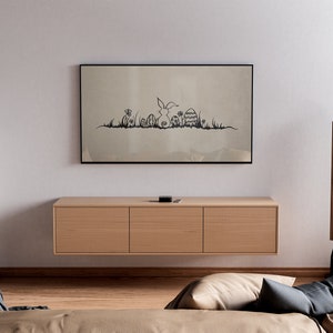 May include: A living room scene featuring a framed artwork with a black and white Easter-themed illustration above a wooden cabinet. Two tall, wooden speakers flank the cabinet. A sofa and armchair are in the foreground.