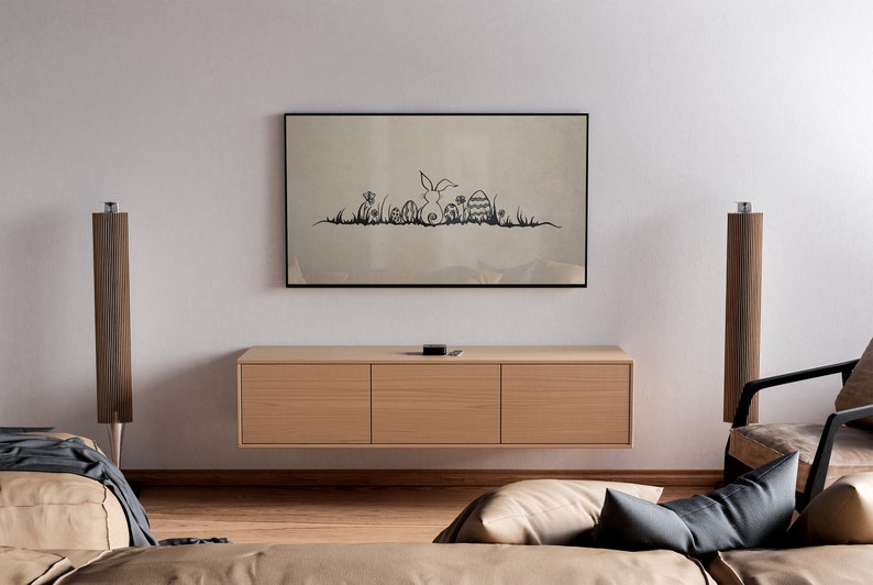 May include: A living room scene featuring a framed artwork with a black and white Easter-themed illustration above a wooden cabinet. Two tall, wooden speakers flank the cabinet. A sofa and armchair are in the foreground.