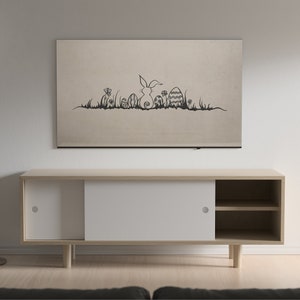 May include: A beige wall-mounted artwork featuring a black line drawing of an Easter bunny, eggs, and grass. Below, a light wood cabinet with white sliding doors and open shelves sits against a white wall. A floor lamp and a gray sofa are also visible.