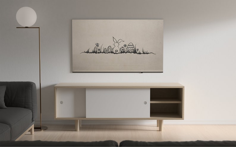 May include: A beige wall-mounted artwork featuring a black line drawing of an Easter bunny, eggs, and grass. Below, a light wood cabinet with white sliding doors and open shelves sits against a white wall. A floor lamp and a gray sofa are also visible.