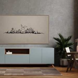May include: A framed artwork featuring a black line drawing of an Easter bunny, eggs, and grass. The artwork is displayed above a light blue cabinet with wooden legs. A potted plant and a small wooden chair are also in the room.