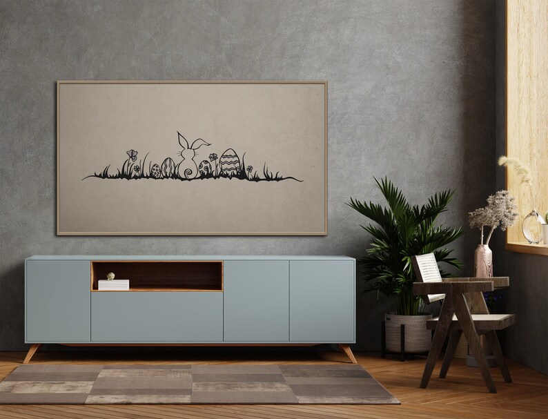 May include: A framed artwork featuring a black line drawing of an Easter bunny, eggs, and grass. The artwork is displayed above a light blue cabinet with wooden legs. A potted plant and a small wooden chair are also in the room.