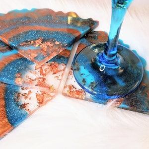 Könnte beinhalten: Satz mit vier Untersetzern aus blauer und kupferfarbener Harz mit goldenen Flocken. Die Untersetzer haben eine wellige Kante und eine glitzernde Oberfläche. Ein blaues Glas steht auf einem der Untersetzer.