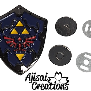 May include: A black metal shield with a blue background and a yellow and red Triforce symbol. The shield is attached to a magnetic clasp with two silver and black metal pieces.