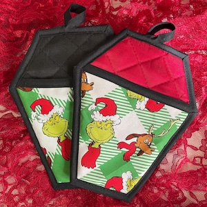 May include: Two quilted pot holders with black trim and hanging loops. One is black, the other has a red top and a green and white plaid pattern with Grinch and Max characters in Santa hats. A festive holiday kitchen item.