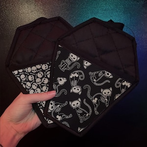 May include: Two black quilted pot holders. One has a white skull pattern, the other has a white skeleton cat pattern. The pot holders are held in a hand.