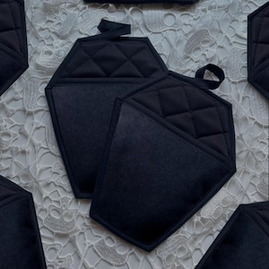 May include: Black quilted hexagon-shaped pot holders with a loop for hanging. The pot holders have a diamond pattern on the top and a solid black pocket. The pot holders are designed to protect hands from heat.