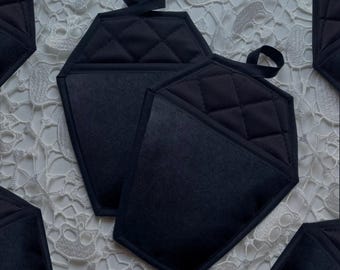 May include: Black quilted hexagon-shaped pot holders with a loop for hanging. The pot holders have a diamond pattern on the top and a solid black pocket. The pot holders are designed to protect hands from heat.