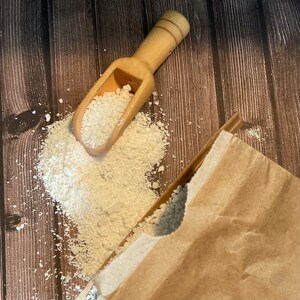 May include: A wooden scoop filled with white powder sits on a brown wooden surface. The powder is spilling out of the scoop and onto the surface. A brown paper bag is partially visible in the background.