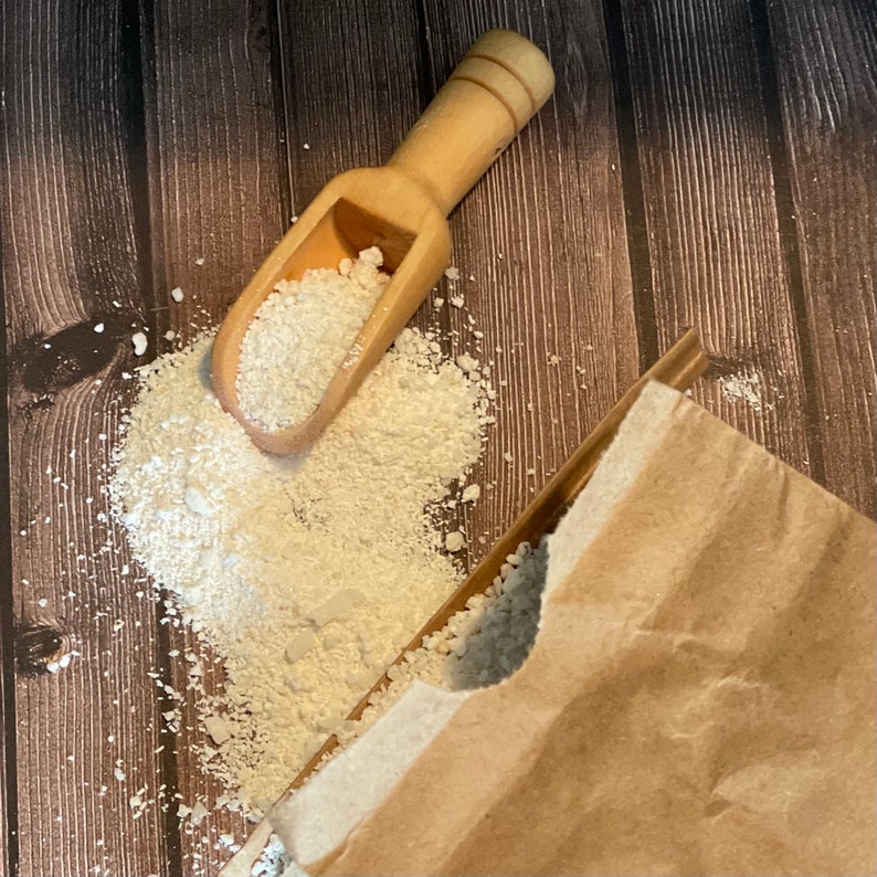 May include: A wooden scoop filled with white powder sits on a brown wooden surface. The powder is spilling out of the scoop and onto the surface. A brown paper bag is partially visible in the background.