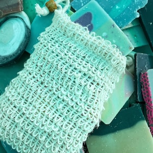 Peut inclure: Un sac en crochet blanc avec un cordon de serrage et une perle en bois, rempli de savons de différentes nuances de vert et de bleu.