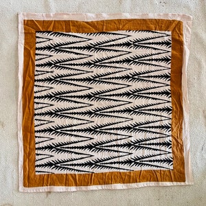 May include: Square textile with a cream-coloured centre featuring a repeating black spiky pattern. The pattern is framed by a mustard-coloured border, which is then framed by a cream-coloured border.