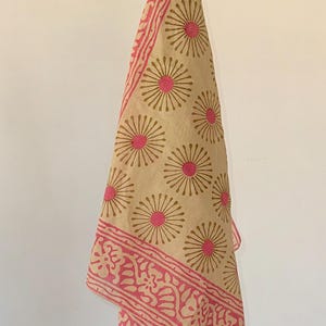 Può includere: Un bandana di cotone beige con un motivo floreale rosa. Il bandana ha un bordo rosa con un motivo floreale. Il bandana è appeso a uno stendino.