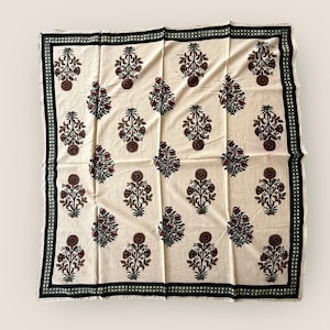May include: Cream-coloured square textile with a repeating floral pattern in burgundy, brown, and green. The border features a black and green dotted design. The fabric is folded, creating a textured appearance.