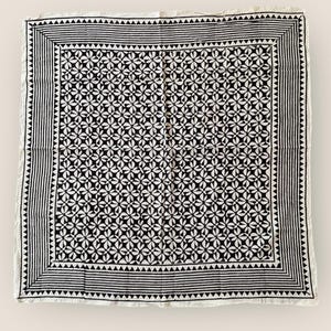 May include: Square black and white patterned textile. The center features a floral design, framed by a border of triangles and stripes. The textile is likely made of cotton or linen.