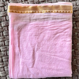 Può includere: Un tessuto di cotone rosa pallido con un bordo dorato. Il tessuto è piegato a metà e ha una consistenza leggermente stropicciata.