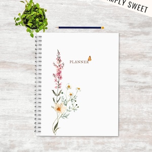 Pode incluir: Uma agenda branca com encadernação em espiral. A capa apresenta uma ilustração em aquarela de flores rosa e brancas, folhas verdes e uma borboleta. O texto "PLANNER" está impresso em marrom na capa.