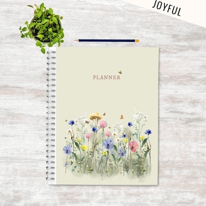 Pode incluir: Uma agenda verde com um design floral com flores rosa, azuis e amarelas. A agenda tem uma encadernação em espiral e a palavra "PLANNER" está impressa na capa.