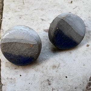 Puede incluir: Dos piedras redondas de cerámica pintadas a mano. Cada piedra está dividida en secciones blancas, grises y azul oscuro. Las piedras están sobre una superficie clara y texturizada.