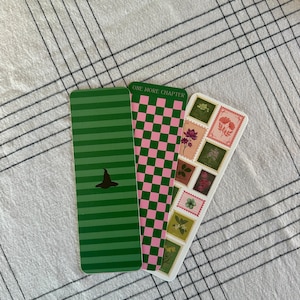 May include: Three rectangular bookmarks. One is green with horizontal stripes and a witch hat silhouette. Another is pink and green checkered. The third has floral postage stamp designs. One bookmark reads "ONE MORE CHAPTER".