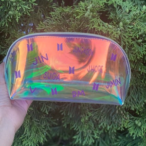 May include: A clear iridescent makeup bag with a zipper closure. The bag is decorated with the names of the members of the South Korean boy band BTS, including Jin, Suga, J-Hope, RM, Jimin, V, and Jungkook. The bag is printed with the BTS logo.