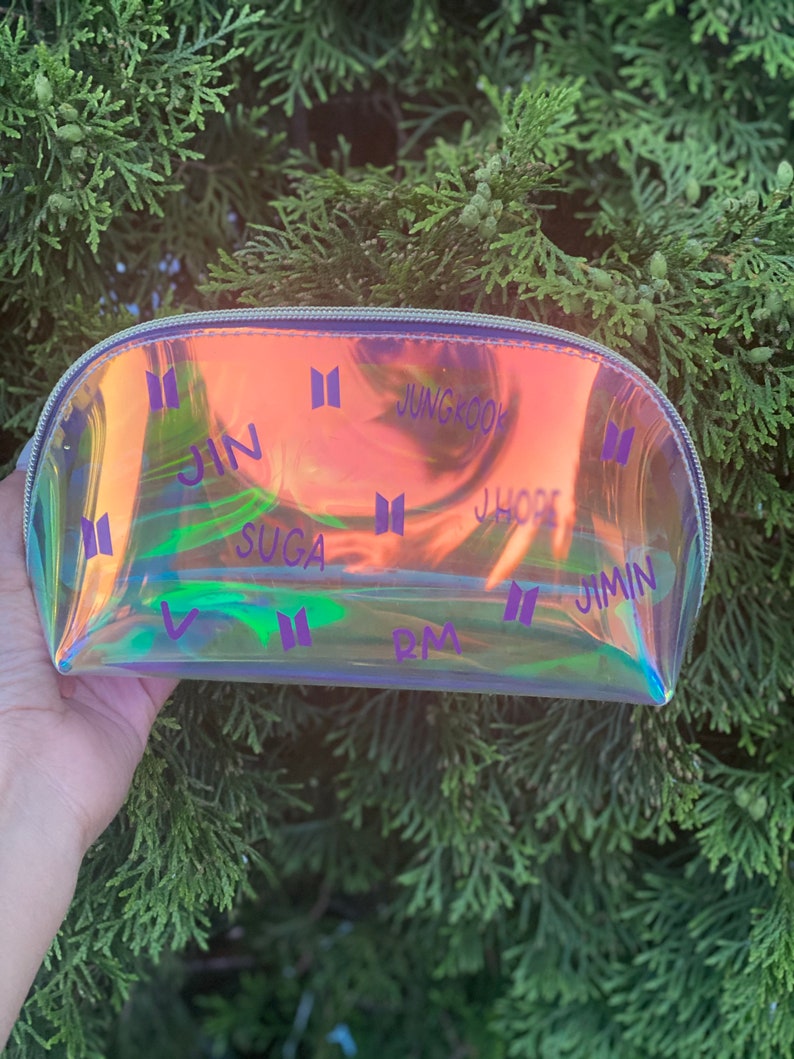 May include: A clear iridescent makeup bag with a zipper closure. The bag is decorated with the names of the members of the South Korean boy band BTS, including Jin, Suga, J-Hope, RM, Jimin, V, and Jungkook. The bag is printed with the BTS logo.