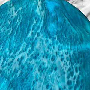 May include: A close-up of a round, ocean-themed resin art piece. The artwork features a blend of turquoise and white, creating a wave-like pattern. The resin has a glossy finish, with visible textures and details.
