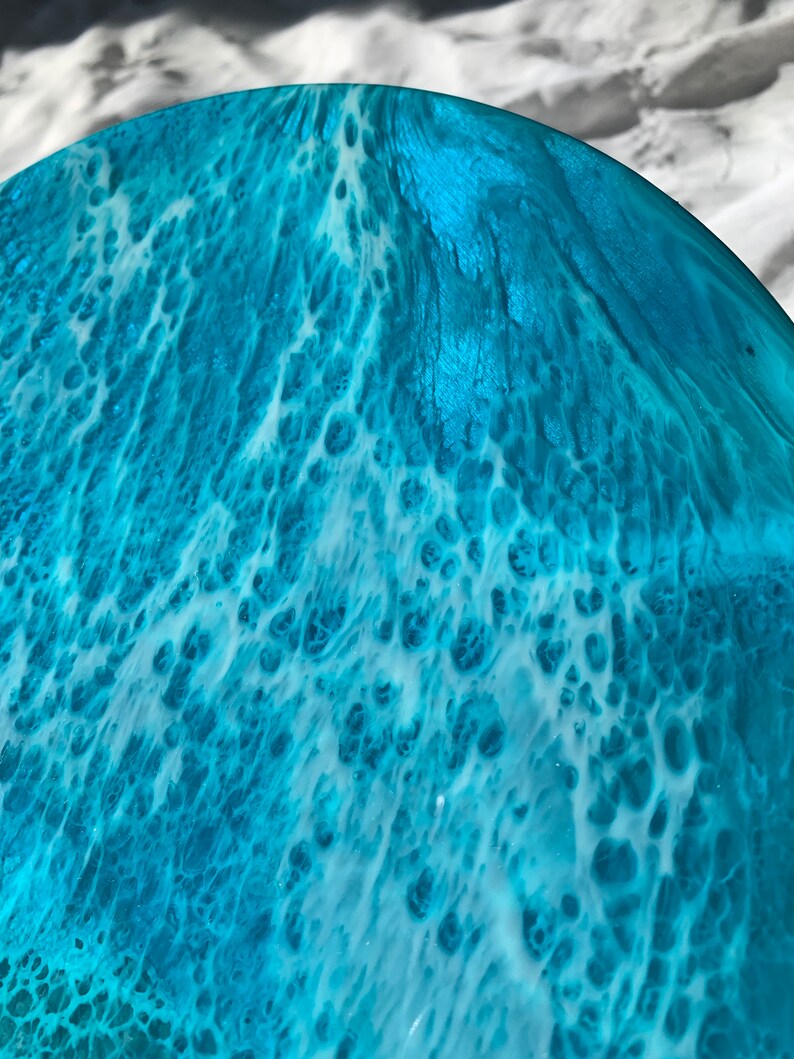 May include: A close-up of a round, ocean-themed resin art piece. The artwork features a blend of turquoise and white, creating a wave-like pattern. The resin has a glossy finish, with visible textures and details.