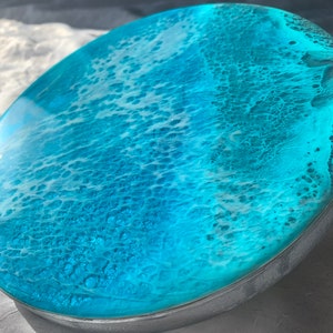 May include: A round, ocean-themed resin art piece with a vibrant turquoise and white design. The artwork features a glossy, wave-like pattern, encased in a silver-colored metal frame. The resin art evokes a coastal theme.