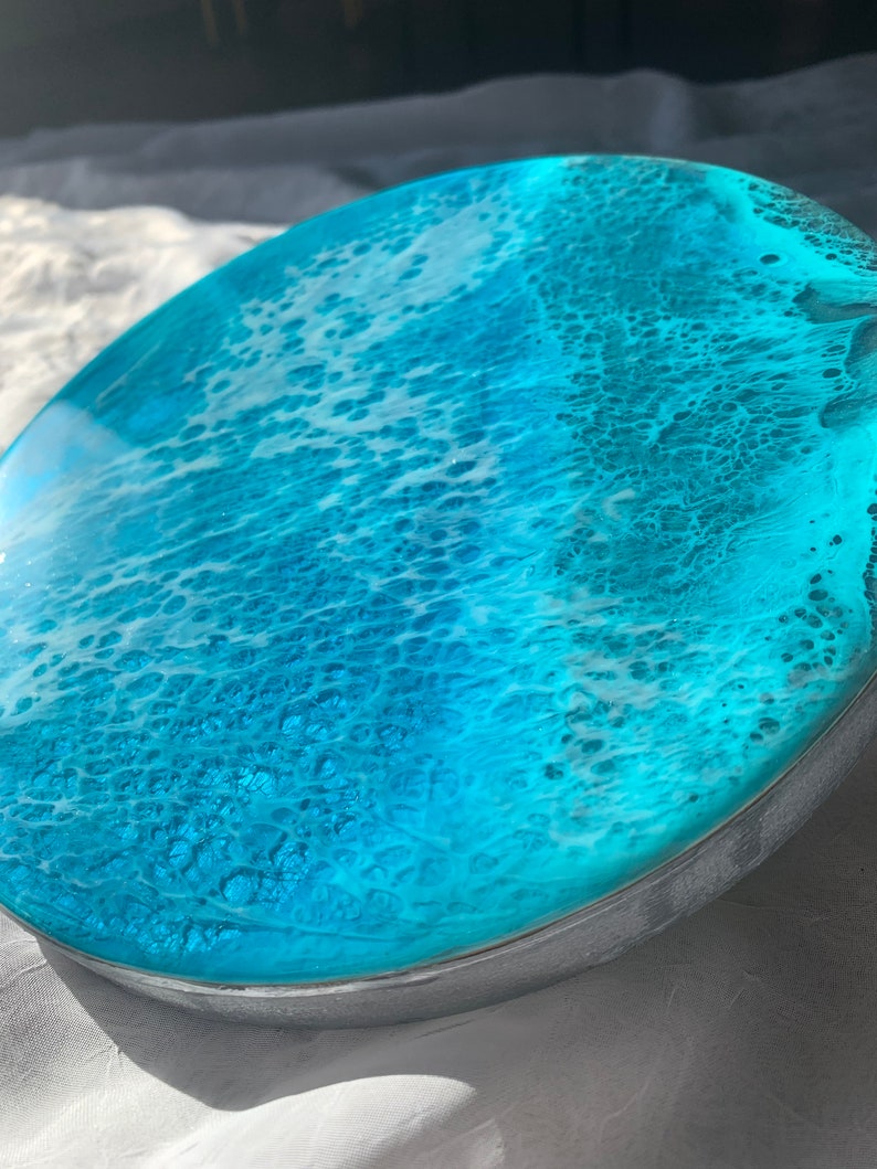 May include: A round, ocean-themed resin art piece with a vibrant turquoise and white design. The artwork features a glossy, wave-like pattern, encased in a silver-colored metal frame. The resin art evokes a coastal theme.