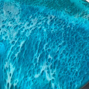 May include: A round decorative tray with a resin ocean wave design. The tray features shades of turquoise and white, mimicking the look of ocean waves. The resin has a glossy finish, creating a textured, three-dimensional effect.