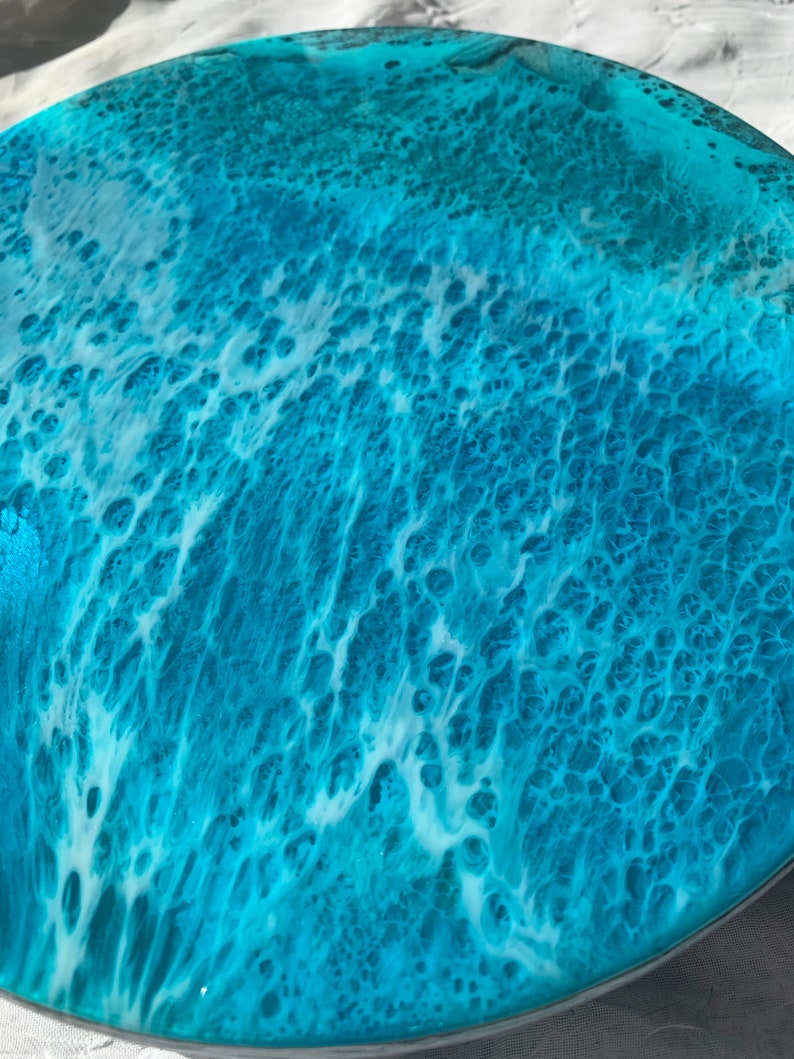 May include: A round decorative tray with a resin ocean wave design. The tray features shades of turquoise and white, mimicking the look of ocean waves. The resin has a glossy finish, creating a textured, three-dimensional effect.