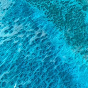 May include: Close-up of a surface with a swirling pattern of turquoise and white resin. The design resembles ocean waves with foamy white crests and deep blue-green depths. The glossy finish reflects light, enhancing the watery effect.
