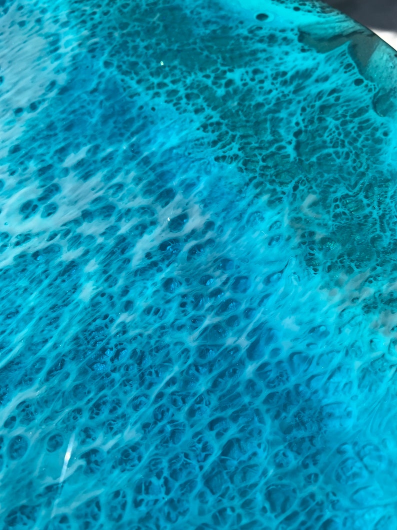 May include: Close-up of a surface with a swirling pattern of turquoise and white resin. The design resembles ocean waves with foamy white crests and deep blue-green depths. The glossy finish reflects light, enhancing the watery effect.