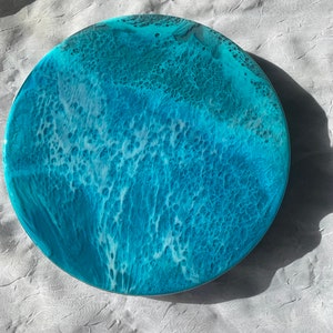May include: Round resin art piece with a coastal theme. The artwork features shades of blue and white, mimicking ocean waves. The circular piece has a glossy finish and a textured appearance, with a diameter of approximately 10 inches.