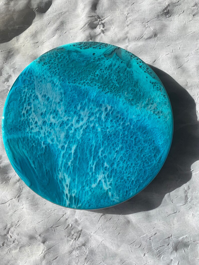 May include: Round resin art piece with a coastal theme. The artwork features shades of blue and white, mimicking ocean waves. The circular piece has a glossy finish and a textured appearance, with a diameter of approximately 10 inches.