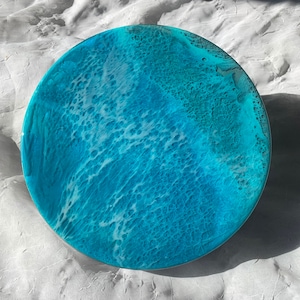 May include: Round resin art piece with a vibrant ocean wave design. The artwork features shades of turquoise and white, creating a dynamic and textured surface. The circular shape is reminiscent of a coaster or decorative plate.