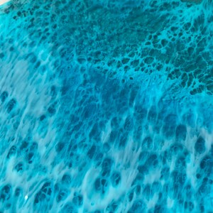 May include: Close-up of a round, resin art piece with a swirling ocean-like design. The artwork features shades of turquoise, teal, and white, creating a textured, wave-like effect. The resin has a glossy finish.