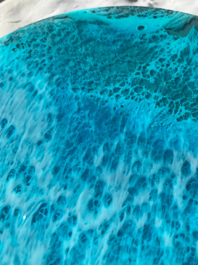 May include: Close-up of a round, resin art piece with a swirling ocean-like design. The artwork features shades of turquoise, teal, and white, creating a textured, wave-like effect. The resin has a glossy finish.