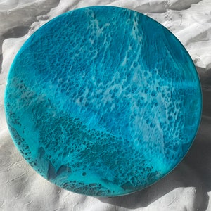 May include: Round resin art piece with a vibrant ocean wave design. The artwork features shades of turquoise, teal, and white, creating a dynamic and textured appearance. The circular shape is approximately 12 inches in diameter.