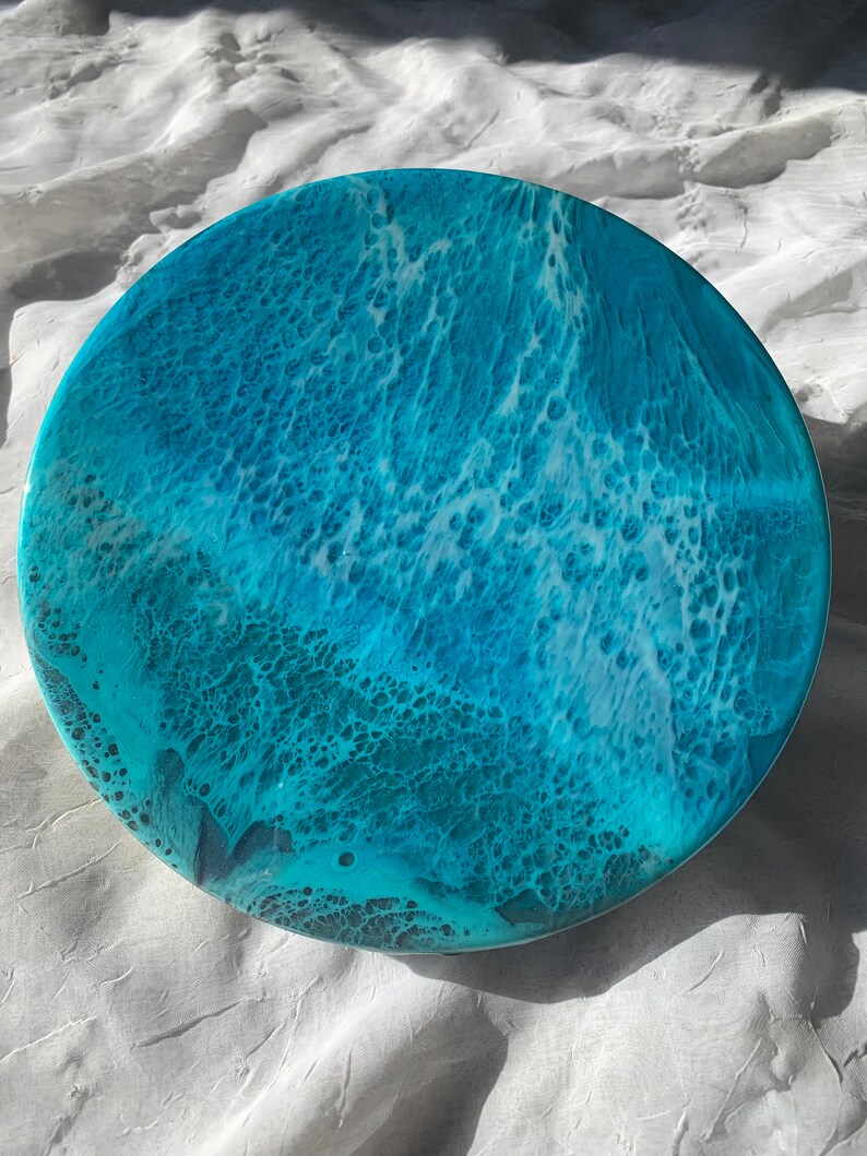May include: Round resin art piece with a vibrant ocean wave design. The artwork features shades of turquoise, teal, and white, creating a dynamic and textured appearance. The circular shape is approximately 12 inches in diameter.